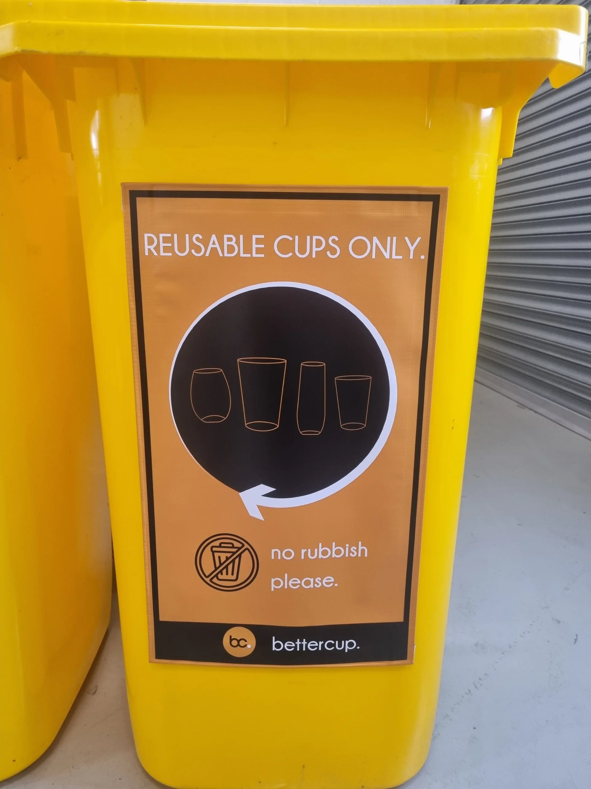 Orange and black sign attached to a yellow wheelie bin that says Reusable Cups Only, with a black circle of cups on it used at festivals and events for collecting cups.