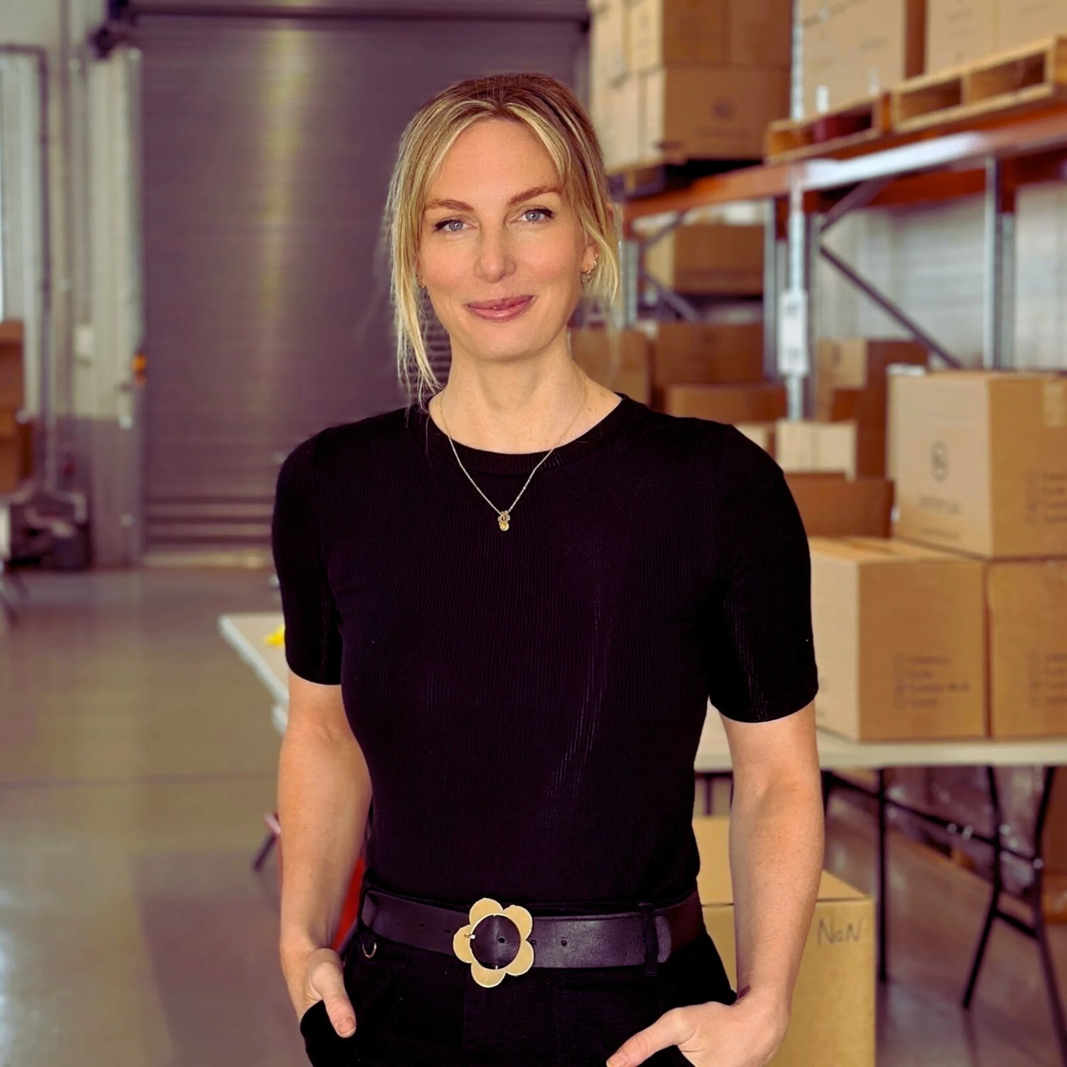 Christie Kamphuis, co-founder of bettercup reuseable cups and reuse systems. A woman with blonde hair wearing a black top and black pants with a flower-shaped belt standing in a warehouse with box of reusable products in a warehouse in australia.