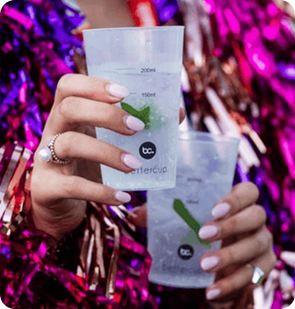 Person holding two bettercup tasters cups with drinks. The person has long white nails, a pearl ring, and is wearing a sparkly, colorful outfit.
