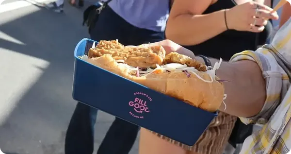 A person holding a Fillgood betterdish with a hot dog topped with fried chicken pieces at an outdoor event, with people waiting in line in the background.