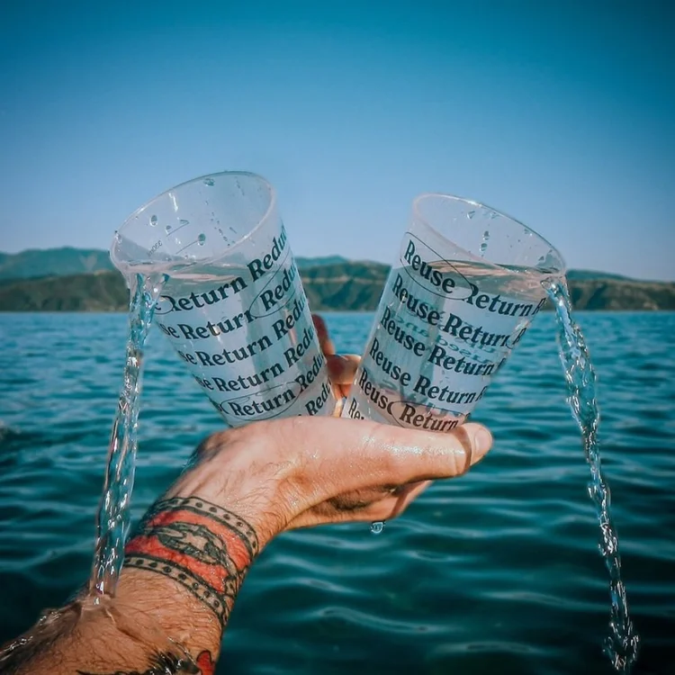 A person with a tattooed arm holding two plastic cups labeled 'Return' and 'Reuse' while pouring water into them against a lake and mountain backdrop.